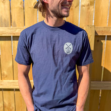 Load image into Gallery viewer, Man wearing a blue t-shirt with a logo against a wooden fence.