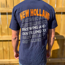 Load image into Gallery viewer, Person wearing a navy blue t-shirt with 'New Holland Brewing and Distilling Co.' text on the back, standing against a wooden fence.
