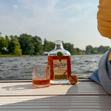 Load image into Gallery viewer, A bottle of The Walley Bunk rum and a matching cut-glass tumbler filled with amber rum, placed on a boat deck with water and trees in the background. A person wearing a yellow hat and blue shirt is partially visible on the right.