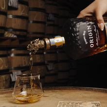 Load image into Gallery viewer, Person pouring whiskey from a bottle labeled 'Origin' into a glass on wooden barrels.

