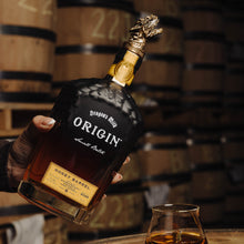 Load image into Gallery viewer, Bottle of Origin Honey Barrel Whiskey held by a hand with a glass on a wooden barrel, barrels in the background.
