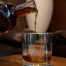 Load image into Gallery viewer, Alcohol being poured from a bottle into a glass with ice cubes on a wooden surface.
