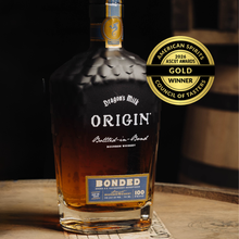 Load image into Gallery viewer, Dragon's Milk Origin Bonded bourbon whiskey bottle with a gold award label on a wooden surface.
