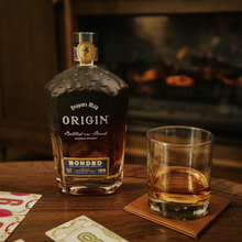 Load image into Gallery viewer, Bottle of Bonded Origin whiskey on a wooden table with a glass filled with whiskey, cards, and a fireplace in the background.
