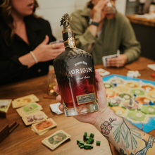 Load image into Gallery viewer, Person holding a bottle of 'Origin' whiskey at a table with a board game
