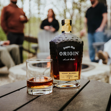 Load image into Gallery viewer, Bottle of 'Origin Honey Barrel Bourbon Whiskey with a glass on a wooden table outdoors, people in the background.
